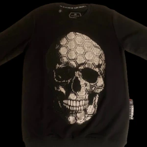 Svart tröja med dödskalle från qp - Snygg svart tröja från Philipp Plein med ett stort dödskallemotiv på framsidan. Tröjan har långa ärmar och en rund halsringning. Perfekt för en cool och edgy stil.
