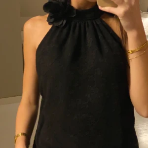 Svart ärmlös blus med blomdetalj - Elegant svart blus med en vacker blomdetalj vid halsen. Den är ärmlös och har en stilren design som passar perfekt för festliga tillfällen. 