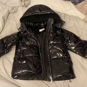 Svart glansig pufferjacka med huva - Säljer en svart glansig pufferjacka från Nelly i storlek XS