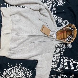 Grå huvtröja från Burberry - Säljer en stilren grå huvtröja från Burberry med dragkedja och klassiskt rutigt foder i huvan. Tröjan har ribbade muddar och en broderad logotyp på bröstet.  Helt oanvänd säljer pga att den är för liten för mig det står att den är M men den passar S