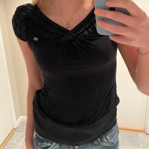 Svart topp från Miss Sixty - Snygg svart topp från Miss Sixty med rynkade detaljer vid halsringningen. Toppen har en v-ringad design och korta ärmar, perfekt för en stilren look. Passar bra till både jeans och kjol. Har även en resår nertill som är samma material som uppe på tröjan. Miss Sixty loggan synligt på bröstet