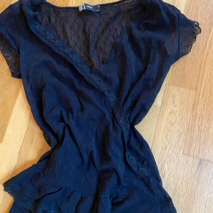 Svart spetsblus från ONLY - Säljer en elegant svart blus från ONLY med spetsdetaljer. Blusen har korta ärmar och en omlott-design som ger en stilren look. Perfekt för att lägga till en touch av elegans i din garderob.