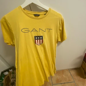 Gul t-shirt från GANT - Snygg gul t-shirt från GANT med tryck av logotyp och ett sköldmärke på bröstet. Perfekt för en avslappnad stil. Kortärmad och i mjukt material.