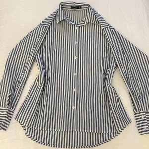 Blå- och vitrandig skjorta - Snygg och stilren skjorta i blått och vitt randigt mönster. Skjortan har långa ärmar och knappar framtill. Perfekt för en klassisk look! Oversize i modellen då den trots storlek XXS sitter oversized på mig som har XS. Tror även att de som har S och M kan ha denna beroende på passformen man vill ha☺️ Endast provad men utan prislapp. 