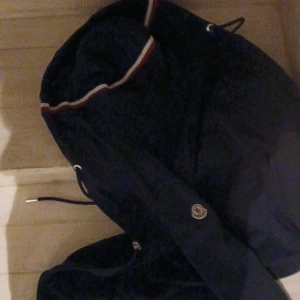 Mörkblå moncler hoodie - Säljer en snygg mörkblå jacka från Moncler med klassiska detaljer. Jackan har en broderad logotyp på ärmen och dragsko med metalländar. Perfekt för kyligare dagar.