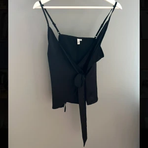 Svart omlottblus från NLY Trend - Svart omlottblus från NLY Trend i storlek 36. Linnet har smala axelband och en knytning i sidan som ger en snygg passform. Perfekt för en kväll ute eller en avslappnad dag. Är i ett glansigare ”siden”-material. Endast använd en gång så är i fint skick! Skicka gärna ett prisförslag om du är intresserad!🖤