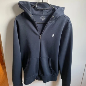Mörkblå hoodie från Polo Ralph Lauren - Säljer en stilren mörkblå hoodie från Polo Ralph Lauren med dragkedja och klassisk logga på bröstet. Tröjan har en bekväm passform med långa ärmar och praktiska fickor framtill. Perfekt för en avslappnad look. 