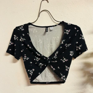 Sexig svart blommig croppad topp från H&M Divided - Säljer en svart croppad topp från H&M Divided med sött blommigt mönster i rosa, rött, vitt och grönt. Toppen har korta ärmar och en knutdetalj framtill som ger en snygg look. Storlek XS. Perfekt för varmare dagar!