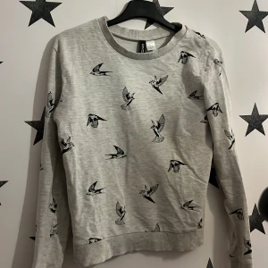 Grå tröja med fågelmönster från H&M Divided - Säljer en grå tröja från H&M Divided med ett coolt fågelmönster i svart. Tröjan har långa ärmar och en rund halsringning. Perfekt för en avslappnad stil. Mer som pyjamas. 
