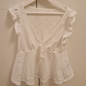 Vit blus - Charmig vit blus med volangärmar. Den har en v-ringad design med knytning framtill. Perfekt för en somrig look.