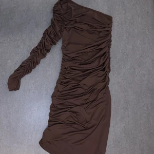 Brun oneshoulder klänning - Snygg brun oneshoulder klänning med rynkade detaljer. Perfekt för en kväll ute eller en speciell tillställning. Den har en lång ärm och en smickrande passform som framhäver figuren.