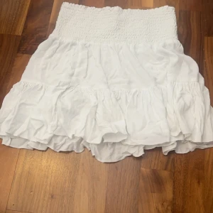 Vit kjol med volangdetaljer - Säljer en söt vit kjol med volangdetaljer och elastisk midja. Har en liten fläck, men man tänker inte på de. Kjolen har en kort längd och är gjord i ett mjukt material.