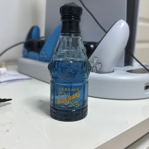 Versace Blue Jeans parfym - Säljer en flaska Versace Blue Jeans parfym. Flaskan är blå och formad som en jeansflaska med detaljerade mönster och text i gult. Perfekt för den som gillar en fräsch och ungdomlig doft.