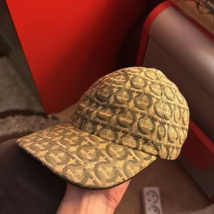 Feragamo keps i bra skick  - Snygg beige feregamo keps med ett unikt mönster och justerbart bälte baktill. Perfekt för att ge din outfit en stilfull touch. Nyligen tvättade och i mycket bra skick. (Ny pris 3,3)