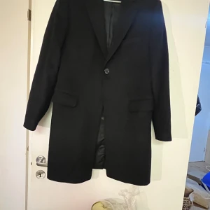 Svart kappa från Cavaliere - Elegant svart kappa från Cavaliere i en klassisk design. Tillverkad av en blandning av ull, viskos och polyester med acetatfoder. Perfekt för en stilren look.