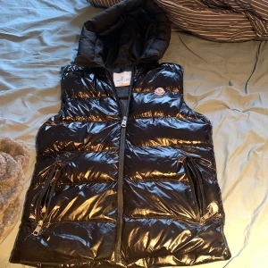 Svart glansig dunväst från Moncler - Snygg svart glansig dunväst från Moncler med dragkedja och huva. Perfekt för kyligare dagar med sin quiltade design. Västen har praktiska fickor med dragkedjor för säker förvaring.