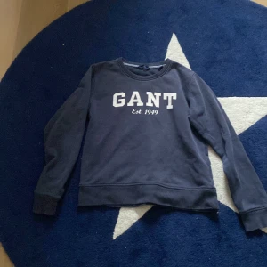 Mörkblå sweatshirt från GANT - Säljer en stilren mörkblå sweatshirt från GANT med vit logotyp på framsidan. Tröjan har långa ärmar och en rund halsringning. Perfekt för en avslappnad stil.