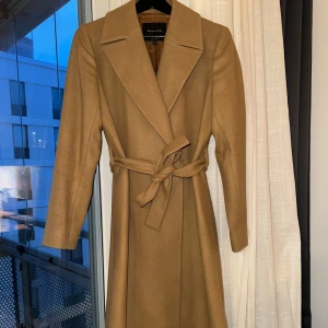 Beige kappa från Massimo Dutti - Elegant beige kappa från Massimo Dutti med klassisk design. Den har en knytning i midjan och långa ärmar. Perfekt för en stilren look under kyligare dagar.