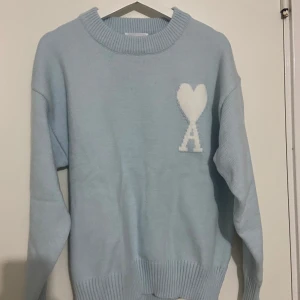 Ljusblå stickad tröja från Ami - Säljer en ljusblå stickad tröja från Ami med ett vitt hjärta och bokstaven A på bröstet. Tröjan har en rund halsringning och långa ärmar. Perfekt för en stilren och mysig look. Den är ganska stor så det funkar både för M och L.