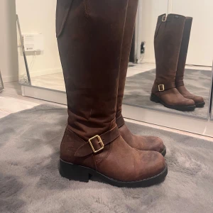 Bruna stövlar med dragkedja och spänne - Snygga bruna stövlar i skinn med dragkedja baktill och dekorativt spänne på sidan. Perfekta för höst och vinter med en robust sula för extra grepp. Stilrena och funktionella för en trendig look.