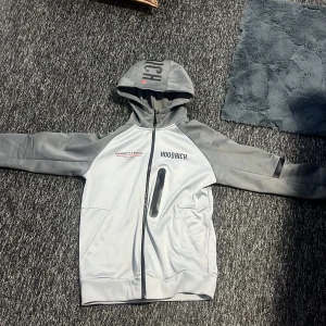 Grå jacka från Hoodrich - Säljer en grå jacka från Hoodrich med huva och dragkedja. Jackan har tryck på både framsidan och baksidan samt en praktisk ficka med dragkedja på bröstet. Perfekt för en casual look.