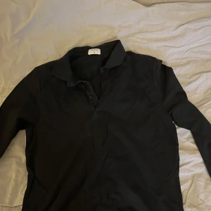 Svart pikétröja från Filippa K - Snygg svart pikétröja från Filippa K med klassisk krage och knappar framtill. Perfekt för en stilren look. Passar bra till både jeans och chinos.