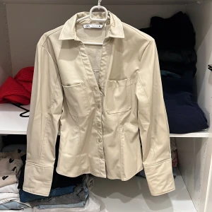 Beige skjorta från Zara - Snygg beige skjorta från Zara med långa ärmar och klassisk krage. Skjortan har en figurnära passform och stilrena fickor framtill. Perfekt för en chic och stilren look.