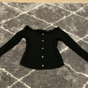 Svart offshoulder topp från Primark - Snygg svart offshoulder topp från Primark med knappar framtill. Toppen har långa ärmar och en ribbad struktur som ger en stilren look. Perfekt för att klä upp eller ner beroende på tillfälle.