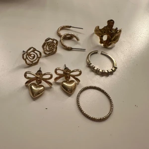 Guldiga örhängen och ringar - Säljer ett set med guldiga smycken som inkluderar örhängen och ringar. Örhängena har olika designer, inklusive rosor och hjärtan med rosetter. Ringarna är enkla och eleganta, perfekta för att komplettera din look.🥰