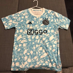 Ajax fotbollströja med blått mönster - Säljer en cool Ajax fotbollströja från Adidas med ett unikt blått och vitt mönster. Tröjan har korta ärmar och klubbens logga på bröstet. Perfekt för fans som vill sticka ut på läktaren eller planen!