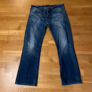 Levis 527 jeans - Säljer ett par klassiska Levis 527 jeans i blå denim. De har en bootcut-stil med slitningar på knäet och en snygg tvättad look. Perfekta för en avslappnad stil. De har några fläckar på vänstra benet som knappt syns (se 4e bilden)