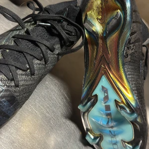 Nike ZM Superfly 10 Elite SE FG - Snygga fotbollsskor från Nike i svart med en cool kromdetalj på sulan. Skorna har snörning och är designade för att ge bra grepp på fasta underlag. Perfekta för den som vill ha stil och funktion på planen.