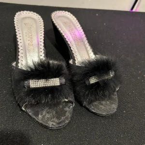 Svarta klackskor med pälsdetalj - Eleganta svarta klackskor från Whisper med en glamorös pälsdetalj och glittrande pärlor på ovansidan. Skorna har en kilklack och öppen tå, vilket ger en lyxig känsla. 