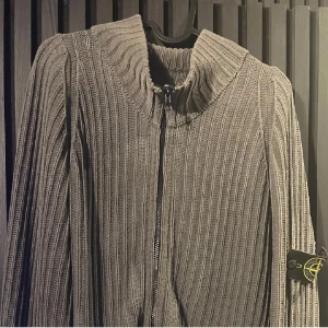 Grå ribbad tröja från Stone Island - Snygg grå ribbad tröja från Stone Island med dragkedja framtill. Tröjan har en hög krage och långa ärmar. Perfekt för en stilren look. M Passar M/L