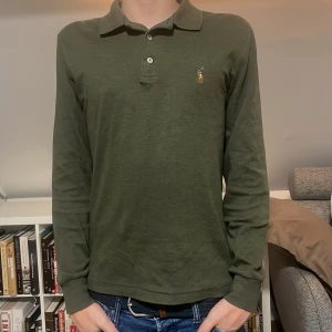 Grön pikétröja från Ralph Lauren - Snygg grön pikétröja från Ralph Lauren med klassisk krage och knappar framtill. Tröjan har långa ärmar och en liten broderad logga på bröstet. Perfekt för en stilren look. Tröjan har en liten fläck men den går bort i tvätten. Perfekt för er som är 165-172 ish. Hör av er vid frågor och funderingar!