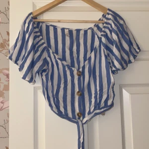 Blå- och vitrandig topp från Amisu - Säljer en superfin blå- och vitrandig topp från Amisu i storlek XL, passar M och L. Toppen har korta ärmar och knappar framtill, samt en söt knytning i nederkant. Perfekt för sommardagar eller en avslappnad vårlook. Tillverkad i bomull vilket gör den skön och luftig att bära.