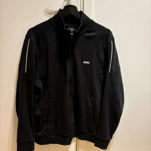 Boss jacka - Säljer en stilren svart jacka från BOSS i mycket bra skick. Perfekt för höst och vår, med en normal passform och långa ärmar. Jackan är gjord i ett mjukt och bekvämt material, perfekt för både vardag och träning. Nypris 2200kr storlek L