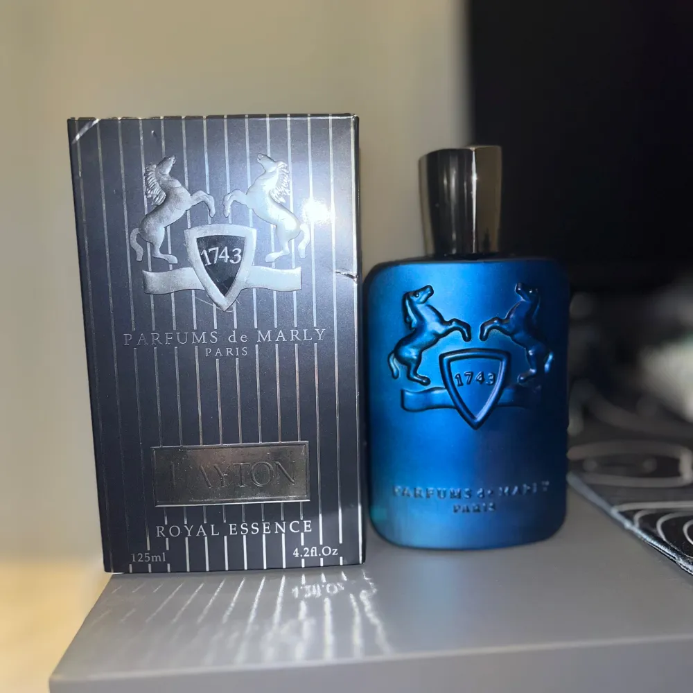 Säljer en lyxig parfym från Parfums de Marly, modellen Layton. Flaskan är elegant i en djupblå färg med en stilren design och ett silverfärgat lock. Förpackningen är svart med silverdetaljer och har en kapacitet på 125 ml. Perfekt för den som vill ha en sofistikerad doft med en touch av elegans.. Parfym.