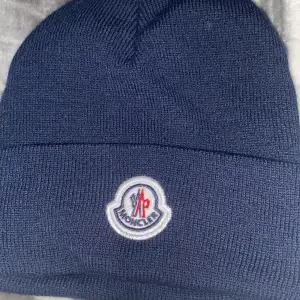 Säljer en snygg mörkblå mössa från Moncler i 100% akryl. Den har en broderad Moncler-logga framtill och är perfekt för kyliga dagar. Mössan är i mycket bra skick och passar perfekt till både vardag och vinteraktiviteter.