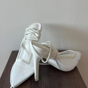 Vita spetsiga klackskor med snörning - Snygga vita klackskor med spetsig tå och elegant snörning. Skorna har en nätliknande design som ger en unik look. Perfekta för festliga tillfällen eller en utekväll. De höga klackarna ger en extra touch av glamour.