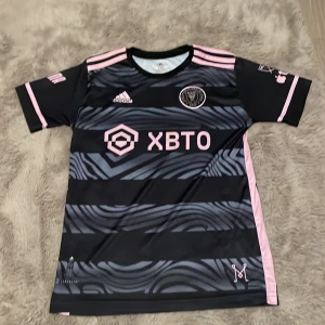 Svart och rosa fotbollströja från Adidas - Säljer en snygg svart och rosa fotbollströja från Adidas. Den har korta ärmar och ett coolt mönster med rosa detaljer. Tröjan är i mycket bra skick och har nummer 10 på ryggen. Perfekt för fotbollsfans! 🌟