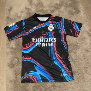 Svart och färgglad fotbollströja från Adidas - Säljer en cool svart fotbollströja från Adidas med färgglada mönster i blått och rött. Tröjan har korta ärmar och är i mycket bra skick. Perfekt för fotbollsfans som vill sticka ut på planen eller läktaren! 🏆