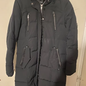 Svart dunjacka från Rockandblue - Säljer en svart dunjacka från Rockandblue i storlek 42. Jackan har en lång modell med dragkedja och flera praktiska fickor. Perfekt för kalla vinterdagar med sin varma och skyddande design. Huvan är avtagbar och ärmarna har dragkedjor för extra stil och funktion.