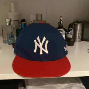 Blå och röd keps från New Era - Säljer en snygg blå keps med röd skärm från New Era. Den har ett broderat vitt NY-logotyp på framsidan och MLB-logotyp på baksidan. Perfekt för sommarens alla äventyr eller för att visa ditt stöd för ditt favoritlag!