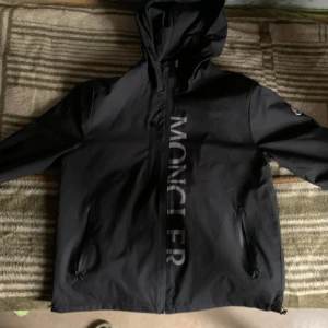 Moncler windbreaker  - Säljer en snygg svart jacka från Moncler i mycket bra skick. Jackan har en dragkedja framtill och en huva för extra skydd. Perfekt för höst och vår, med Moncler-loggan tryckt längs dragkedjan. Den är långärmad och har en stilren design.