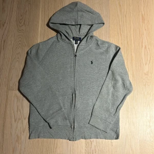 Grå hoodie från Ralph Lauren - Säljer en snygg grå hoodie från Ralph Lauren i storlek L(14-16 år. Den är i bra skick och har en dragkedja framtill. Perfekt för höst och vår, med en klassisk design och det ikoniska logotypbroderiet på bröstet. Superbekväm och stilren!