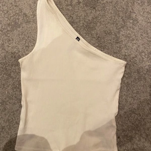 Vit oneshoulder topp från Pieces - Fin vit oneshoulder topp från Pieces i ribbat material. Perfekt för sommardagar eller en utekväll. Toppen har en stilren design och är i mycket bra skick. Endast använd en gång på skolavslutning☺️