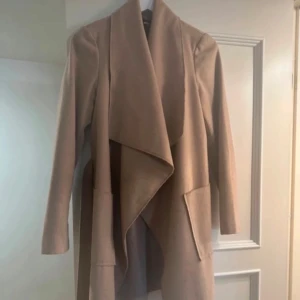 Beige kappa med knytband - Säljer en superfin beige kappa i mycket bra skick. Den har en elegant draperad krage och ett knytband i midjan för en snygg passform. Perfekt för hösten! Nypris runt 900