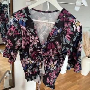 Blommig topp med knytning - Säljer en superfin blommig topp med v-ringning och knytning framtill. Den har trekvartsärmar och ett färgglatt mönster i rosa, lila och blått på svart botten. Perfekt för vår och sommar! 🌸