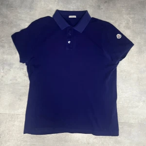Mörkblå pikétröja från Moncler - Säljer en snygg mörkblå pikétröja från Moncler. Den har en klassisk krage med två knappar och Monclers logga på ärmen. Perfekt för både vardag och lite finare tillfällen. Tröjan är i mycket bra skick och passar perfekt till jeans eller chinos. finns en liten fläck på baksidan som man knappast märker Storlek L men passar M